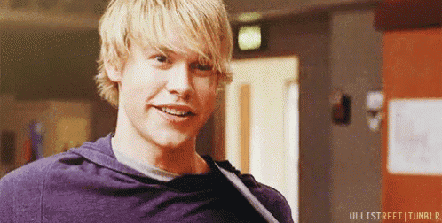 a young man with blonde hair is wearing a purple shirt and smiling in a hallway .