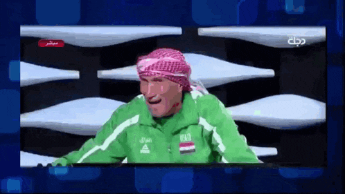 a man wearing a green jacket and a head scarf is laughing on a television screen .