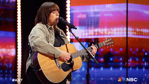 a woman singing into a microphone while holding an acoustic guitar in front of a nbc logo