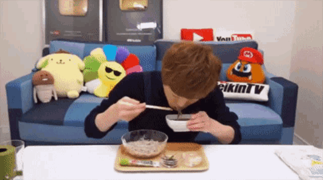 a man is eating from a bowl with chopsticks in front of a couch with a bikinitv pillow on it