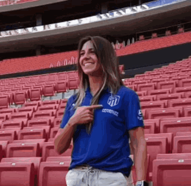 a woman in a blue shirt is standing in an empty stadium pointing at herself .