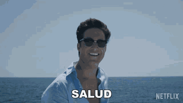 a man wearing sunglasses smiles in front of the ocean and the word salud is above him