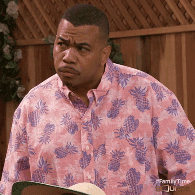 a man wearing a pink shirt with pineapples on it is holding a hat