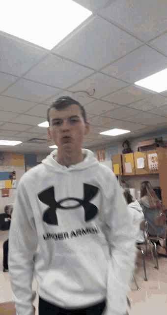 a boy wearing a white under armour sweatshirt stands in a classroom