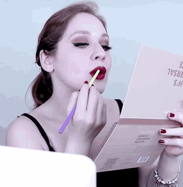 a woman is applying lipstick to her lips while looking at a book that says adam 's makeup palette