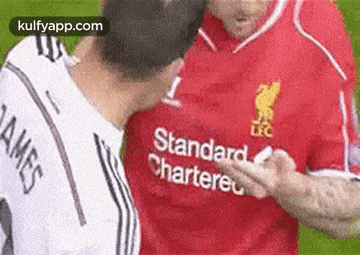 two men are standing next to each other on a soccer field . one of the men is wearing a red standard chartered jersey .