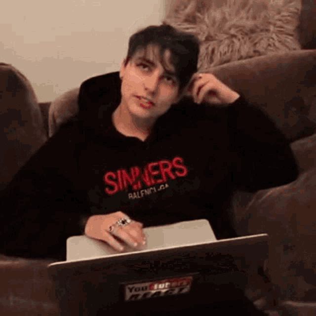 a person is sitting on a couch with a laptop and a hoodie that says sinners on it .