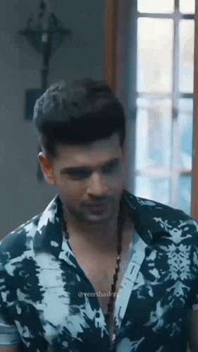 a man in a tie dye shirt and necklace is standing in front of a window .