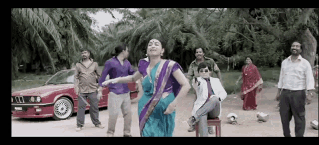 a woman in a blue and purple sari is dancing in front of a red car