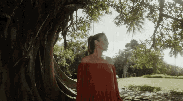 a woman in a red off the shoulder dress stands in front of a tree