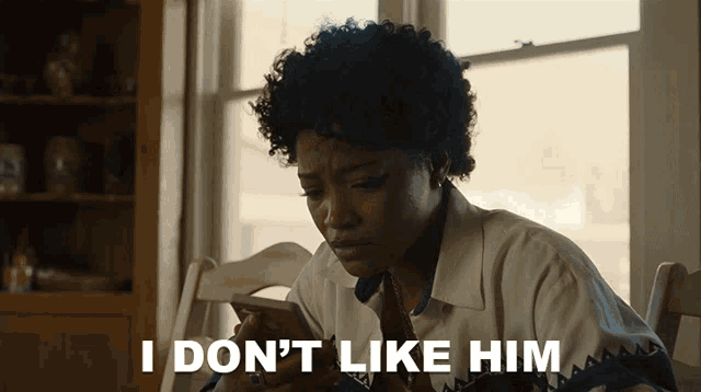 a woman sitting at a table looking at her phone with the words i don 't like him behind her