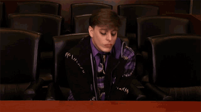 a man in a purple shirt and tie sits in a row of black chairs