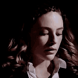 a close up of a woman 's face in a dark room . she is wearing a white shirt and a black jacket .