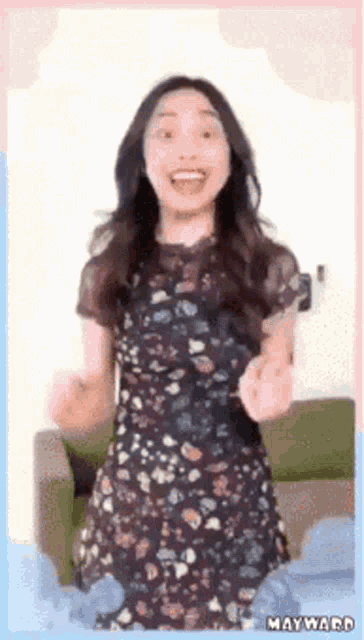 a woman in a floral dress is standing in front of a couch with her arms in the air .