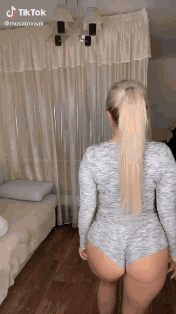 a woman in a grey bodysuit stands in front of a white curtain with tiktok written on the bottom of the screen