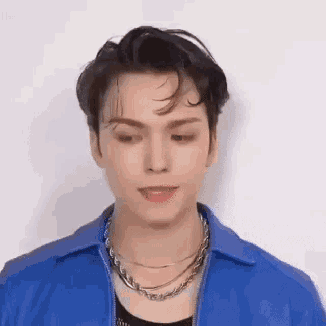 a young man wearing a blue jacket and a necklace is standing in front of a white wall .