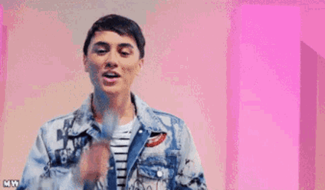 a man in a denim jacket is singing into a microphone with a pink background