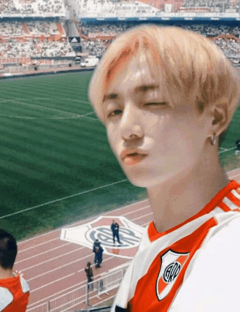 a young man wearing a river plate jersey stands in front of a soccer field with his eyes closed