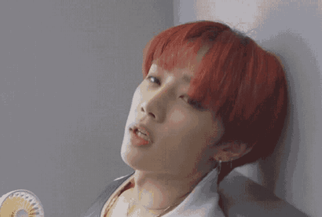 a young man with red hair is leaning against a wall with a fan in his hand .