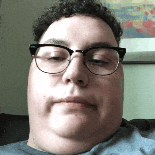 a man with curly hair and glasses looks at the camera with his eyes closed