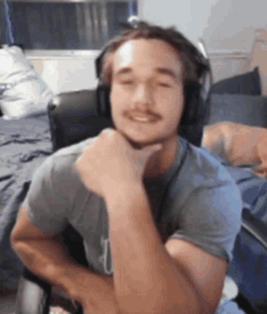 a man wearing headphones and a gray shirt is sitting in a chair with his hand on his chin .
