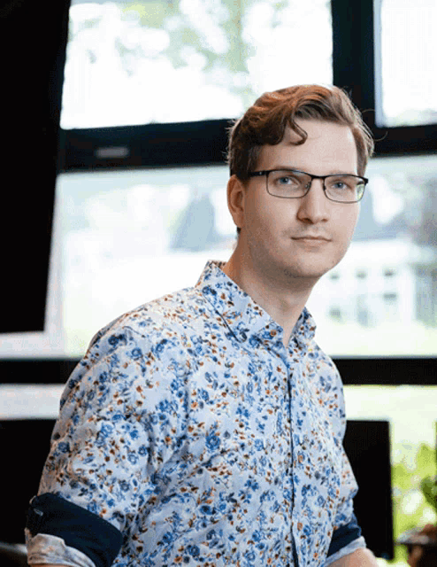 a young man wearing glasses and a floral shirt looks at the camera