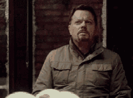 a man with a beard is sitting in front of a brick wall looking at something .