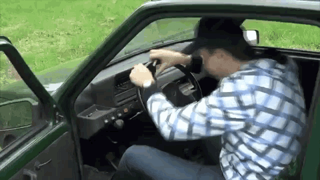 a man in a plaid shirt is driving a green car and adjusting the steering wheel .