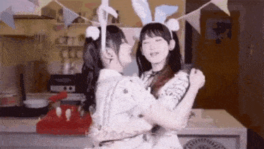 two girls are hugging each other in a kitchen . one of the girls is wearing bunny ears .