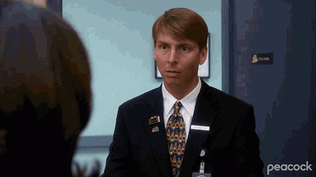 a man in a suit and tie with a peacock logo on the bottom left