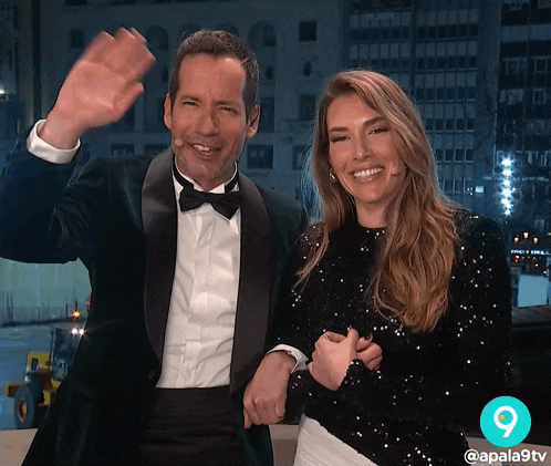a man in a tuxedo and a woman in a sequined top are smiling and waving