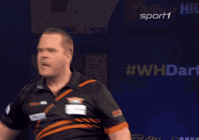 a man in a black and orange shirt is pointing up in front of a screen that says sport1