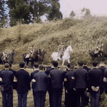 a group of men are standing in a field watching a man riding a white horse