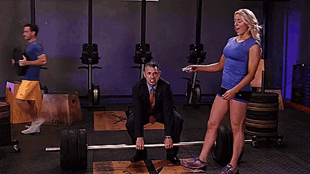 a man is squatting on a barbell in a gym while a woman stands behind him .