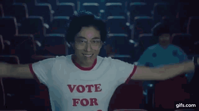 a man wearing a vote for shirt is making a heart shape with his hands in a theater .