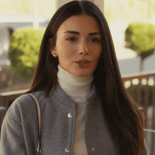 a woman wearing a grey jacket and a white turtleneck looks at the camera